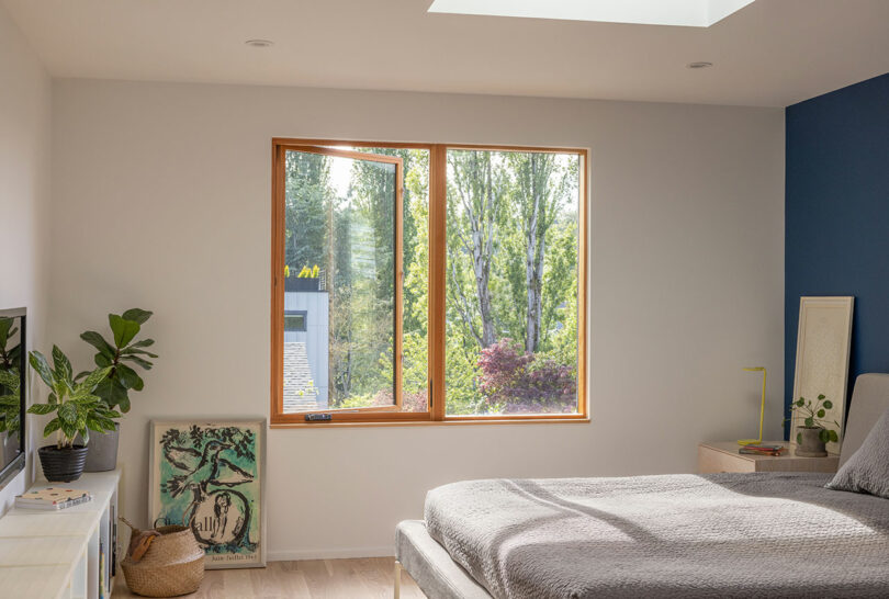 A sunlit bedroom with a large window showing trees outside, a bed with gray bedding, a plant, TV, and artwork leaning against the wall.