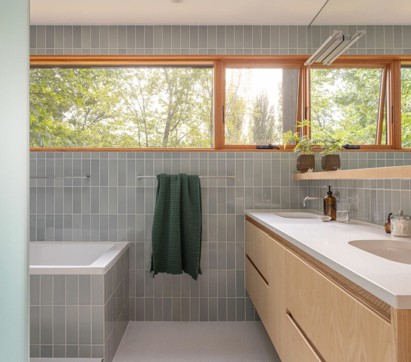 Modern bathroom with light gray tile walls, a built-in bathtub, wooden vanity with dual sinks, large horizontal window, and green towel hanging on a rail.