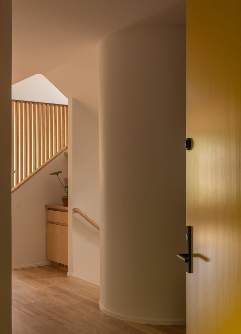 A curved white wall and wooden floor hallway with a yellow door ajar, wooden staircase railing, and a small plant on a cabinet near the stairs.