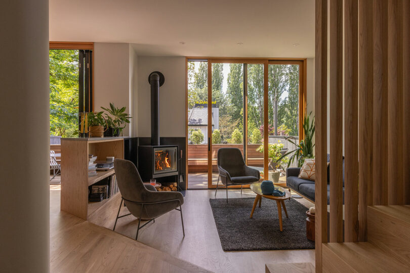 Modern living room with large windows, a wood-burning stove, two armchairs, a sofa, plants, and a view of an outdoor deck with greenery. Natural light fills the space.