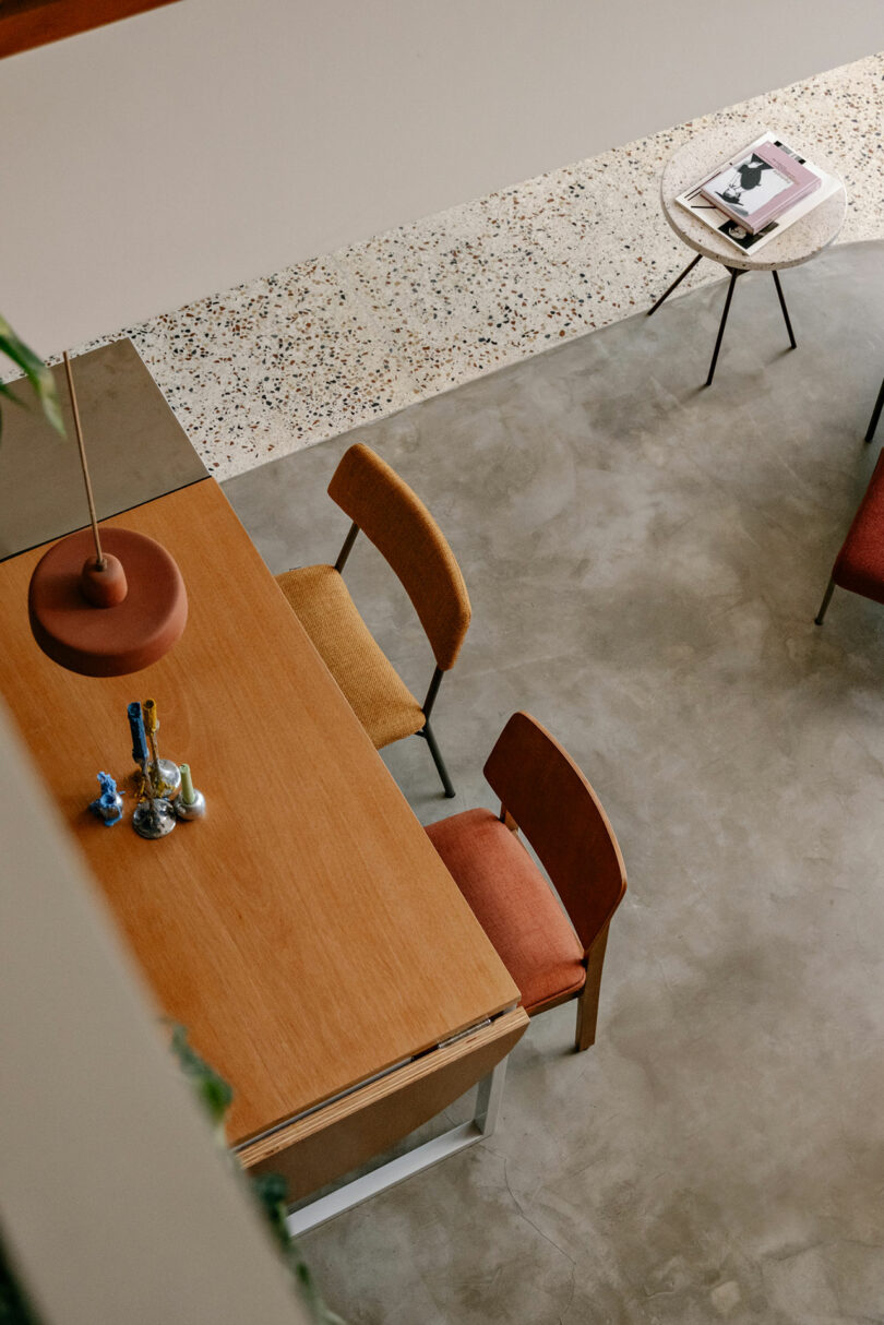 A modern dining table with two wooden chairs, decorative items on the table, and a small side table with magazines on a terrazzo and concrete floor.