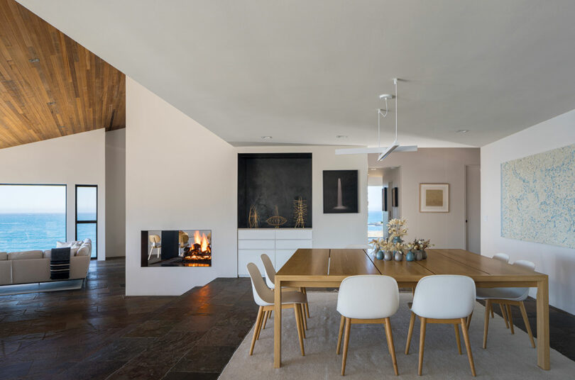 Modern open-plan dining area with a wooden table, white chairs, a built-in fireplace, and ocean view through large windows. Minimalist decor with abstract art on the walls.