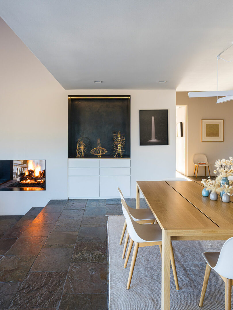Modern dining room with a wooden table, white chairs, decorative items on a shelf, stone tile flooring, and a lit fireplace on the left.