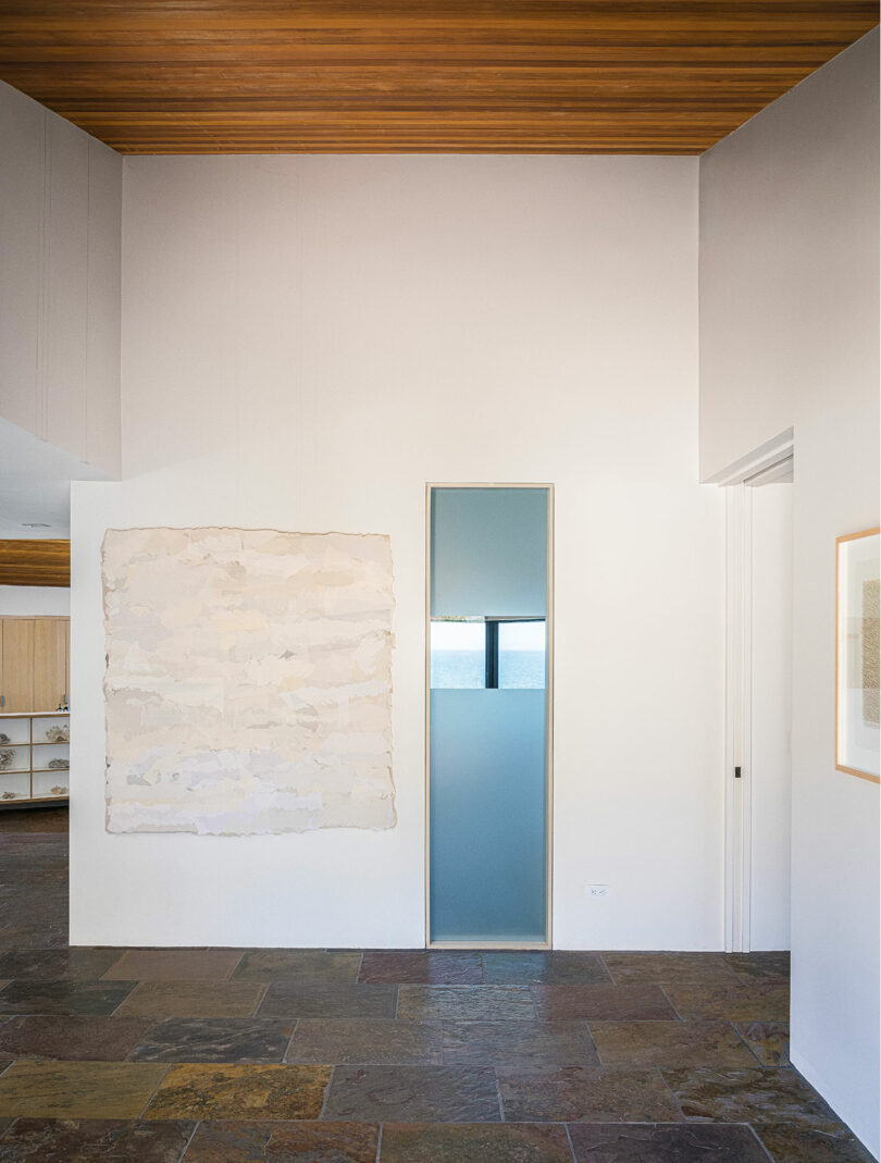 A modern interior with tall white walls, a narrow frosted glass door, slate tile flooring, a wood ceiling, and minimalist artwork.