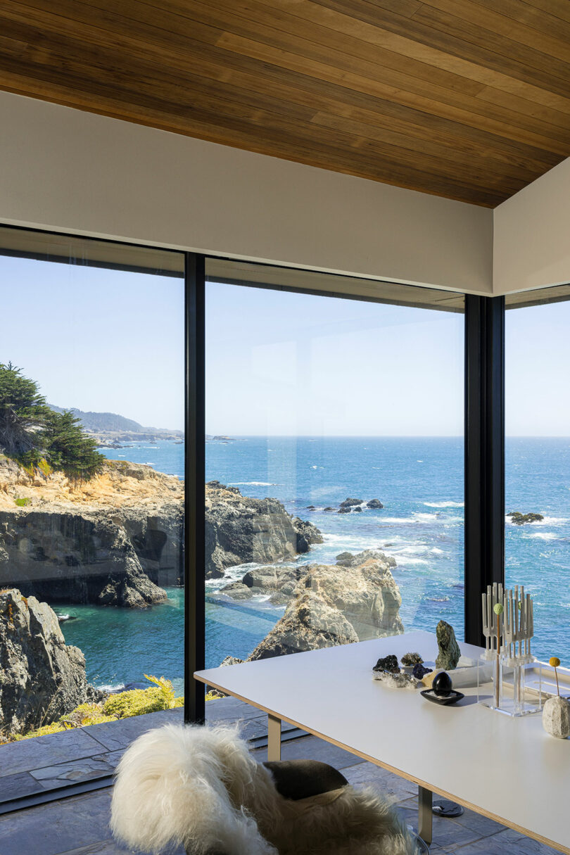 Modern interior with a table and chair next to large floor-to-ceiling windows overlooking a rocky ocean coastline under a clear sky.