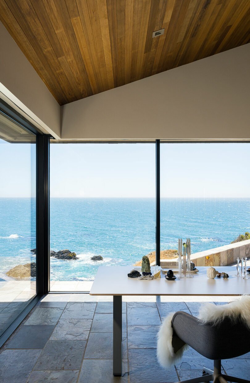 Modern workspace with a large white desk and chair facing floor-to-ceiling windows overlooking the ocean and rocky shoreline.