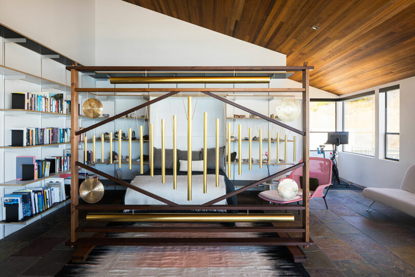 A modern bedroom with a bed separated by a decorative wooden and brass divider, bookshelves on the left, and seating by large windows on the right.
