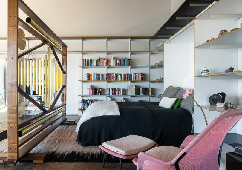 Modern bedroom with open bookshelves, a bed with black bedding, a pink lounge chair, decorative objects, and a wooden divider separating the sleeping area from the rest of the room.