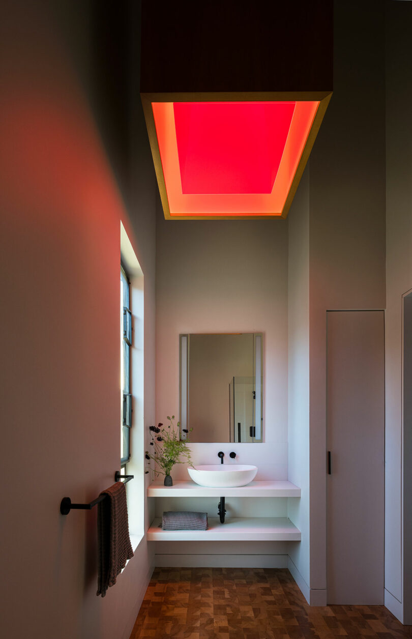 A modern bathroom with a floating white sink, rectangular mirror, and a large ceiling light panel glowing red. A towel hangs on the left, and a window lets in natural light.