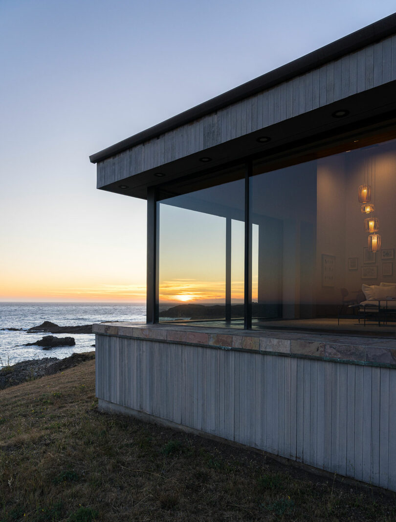 Modern house with large glass windows overlooks the ocean at sunset, with warm interior lights visible and grassy ground in the foreground.