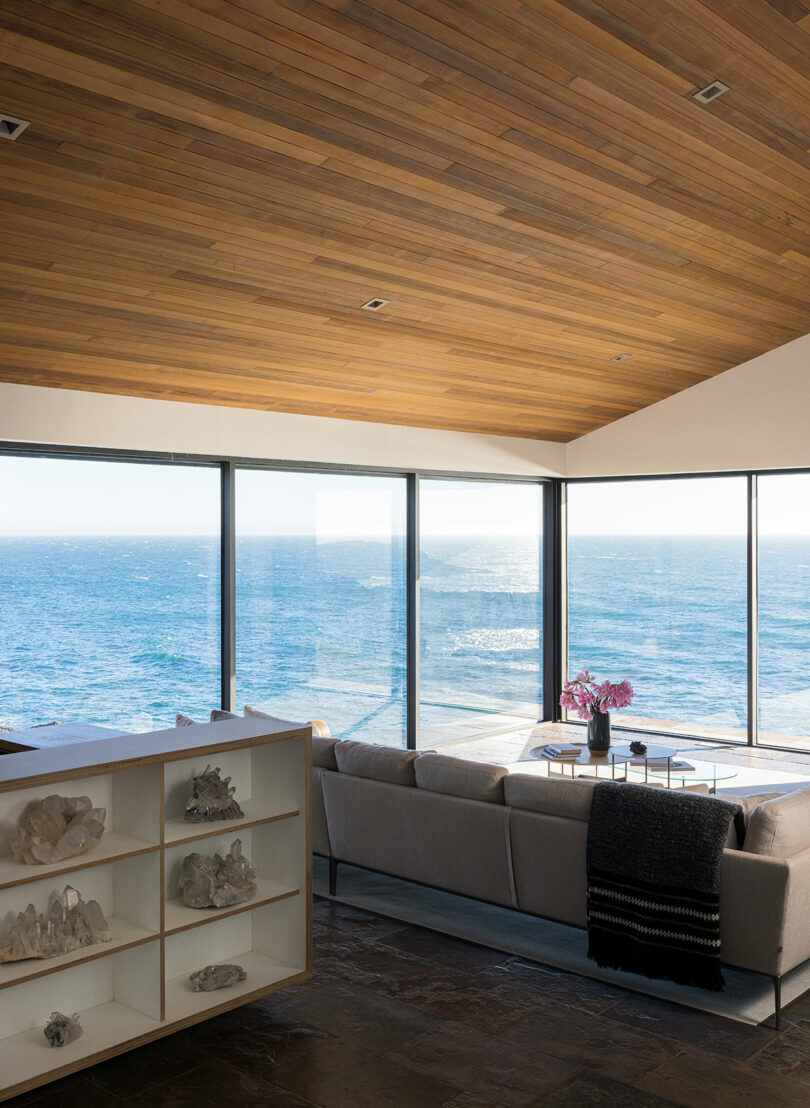Modern living room with floor-to-ceiling windows overlooking the ocean, featuring a light gray sofa, coffee table with flowers, and a display shelf with crystals.