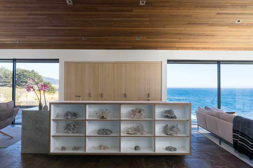 Modern living room with wooden ceiling, open shelves displaying coral, large windows overlooking the ocean, and minimalist furniture.