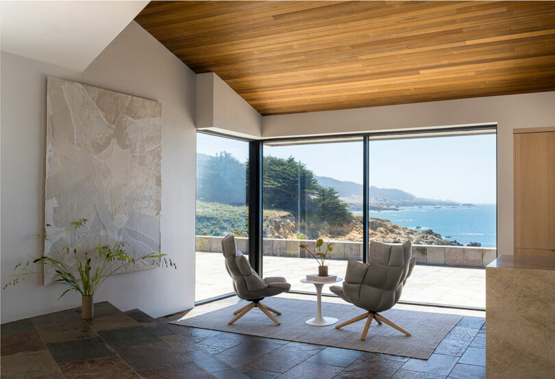Modern sitting area with two armchairs and a small table by large windows, overlooking a coastal view with trees and mountains in the background.