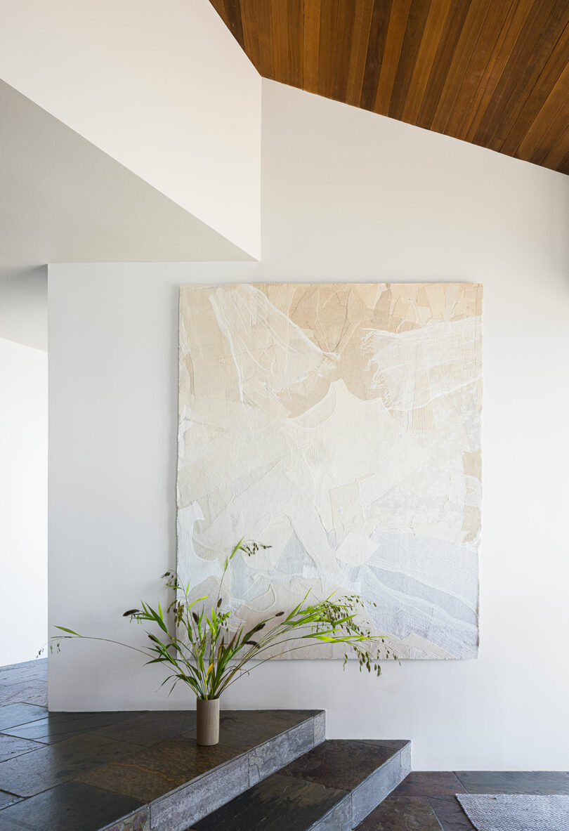 A large abstract beige and white wall hanging is displayed above stone steps with a vase of green branches placed at the base.