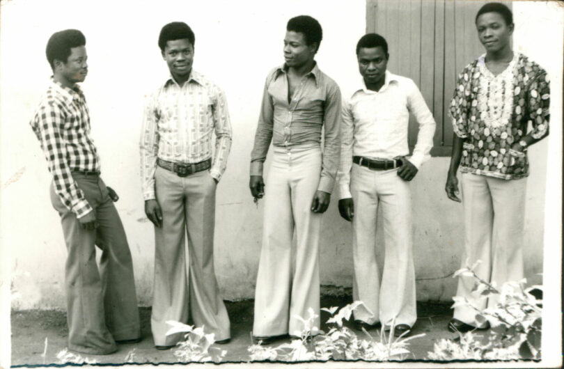 Five men stand in a row against a wall, wearing patterned shirts and flared trousers, looking in different directions.
