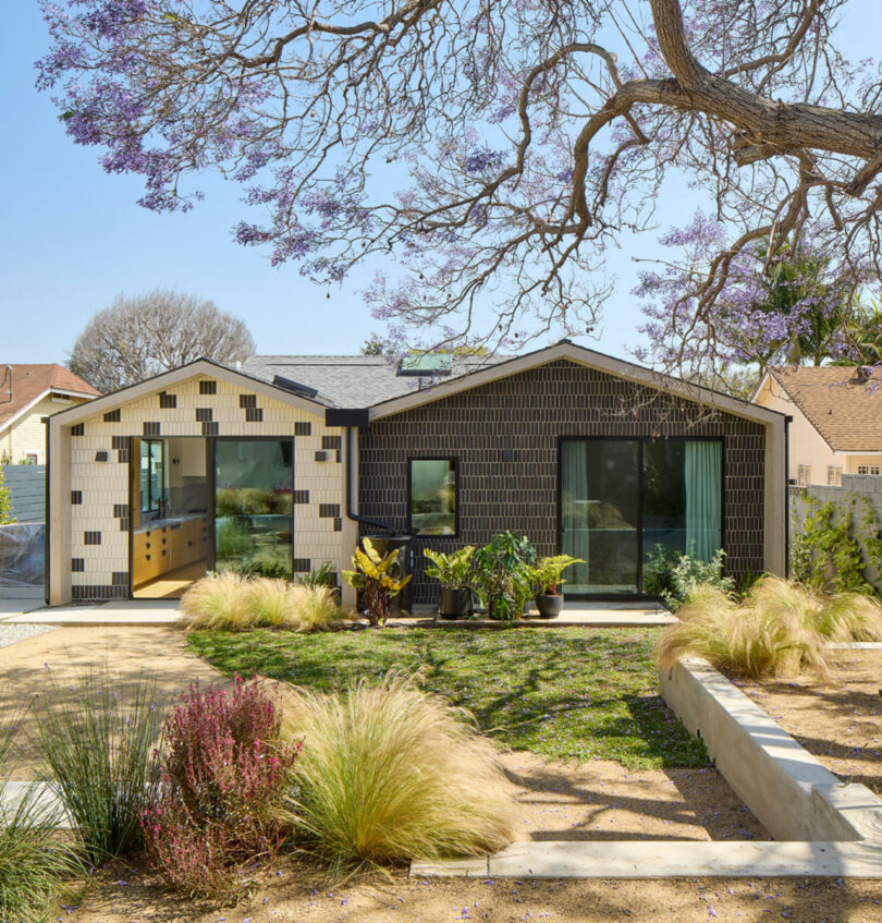 Modern single-story house with black and white geometric exterior, large windows, landscaped front yard, and a tree with purple flowers overhead.