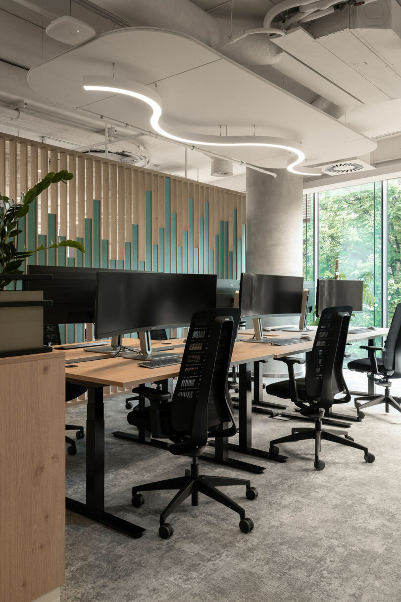 Modern open-plan office by BIT CREATIVE with several empty desks, ergonomic chairs, computer monitors, large windows, and contemporary lighting. Vertical blue wall accents and greenery add a refreshing touch.