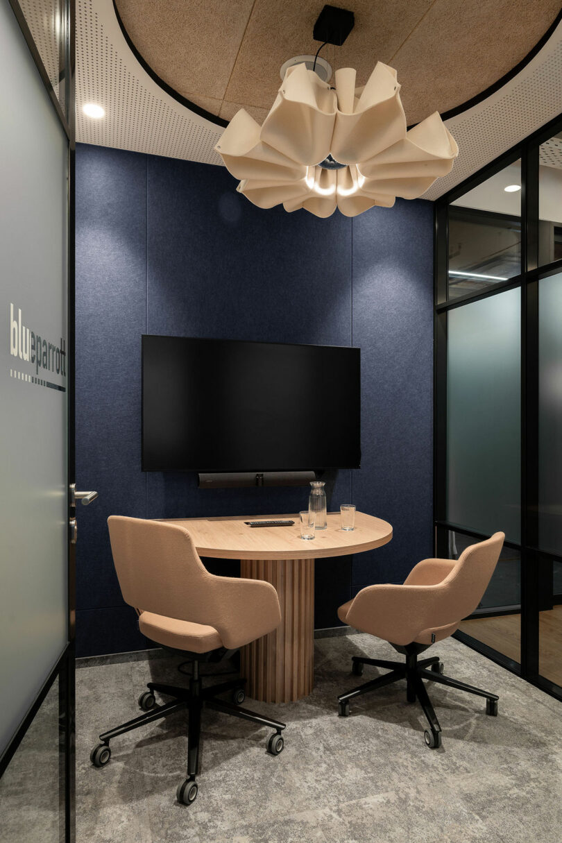A small, modern BIT CREATIVE meeting room with two beige office chairs, a round wooden table, a wall-mounted TV, and a decorative ceiling light fixture.