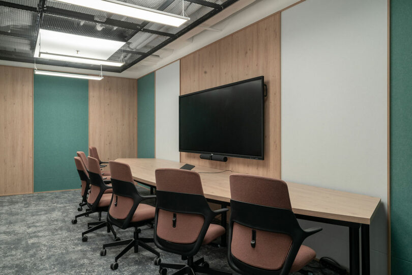 Modern conference room by BIT CREATIVE with a long table, six pink office chairs, a large wall-mounted screen, and minimalist decor featuring wood paneling and green accents.