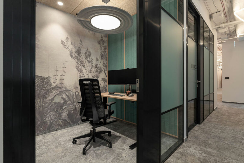 A modern office workspace by BIT CREATIVE features a black ergonomic chair, a desk with a monitor, and stylish wall art, all visible through a glass door from the hallway.