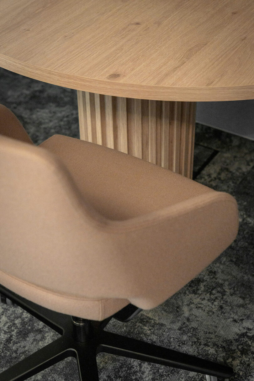 A BIT CREATIVE touch shines in this setup: a beige upholstered office chair sits beside a round wooden table with a ribbed central base, all arranged on a textured gray carpet.