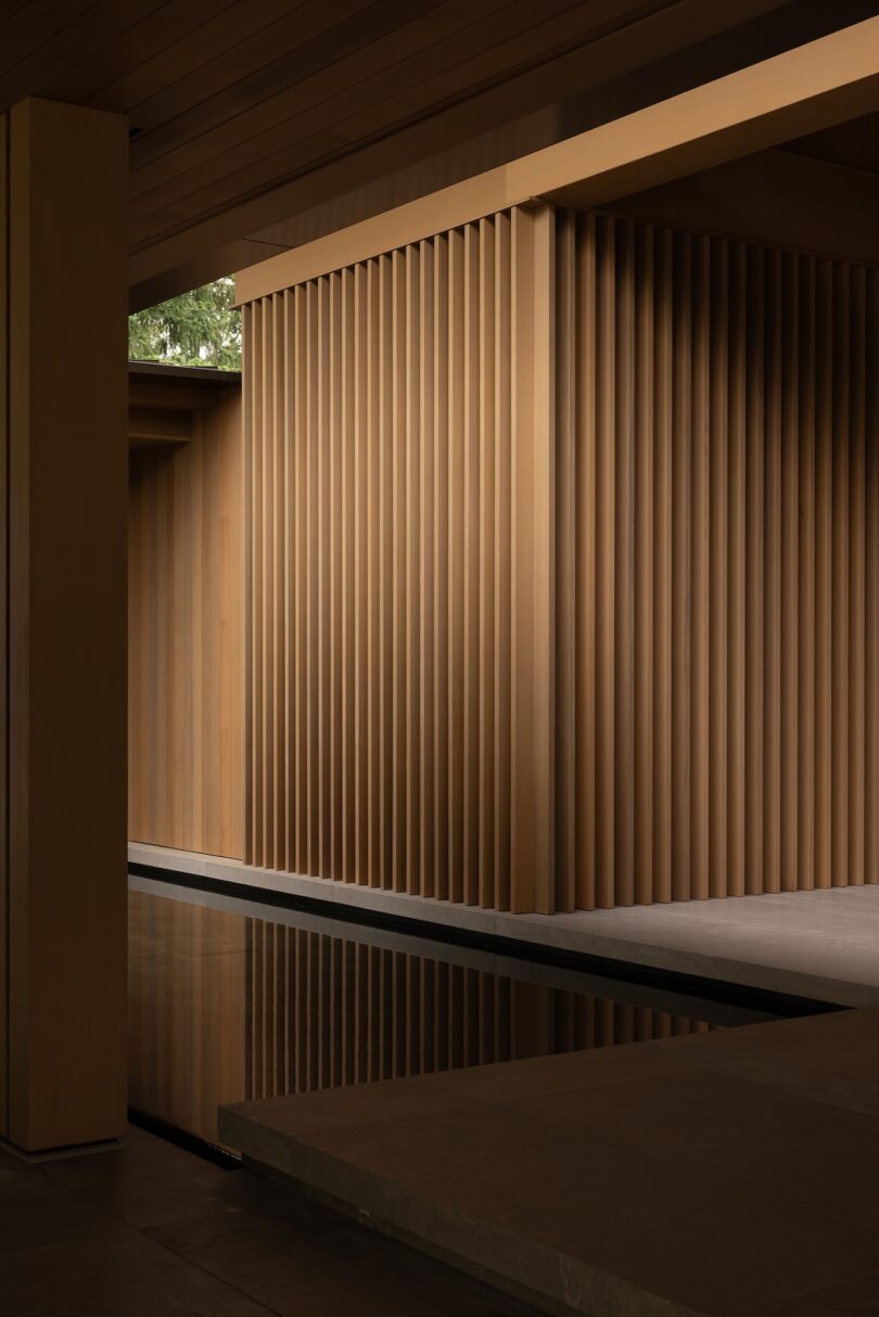 Interior space featuring vertical wooden slats, angled walls, and a shallow reflecting pool, with natural light entering from an opening above.
