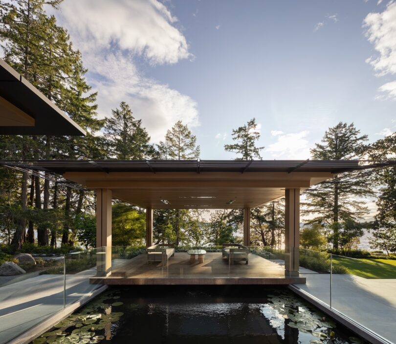 Modern open-air pavilion with wooden roof, glass railings, and outdoor seating, situated above a pond in a landscaped forest setting under a partly cloudy sky.