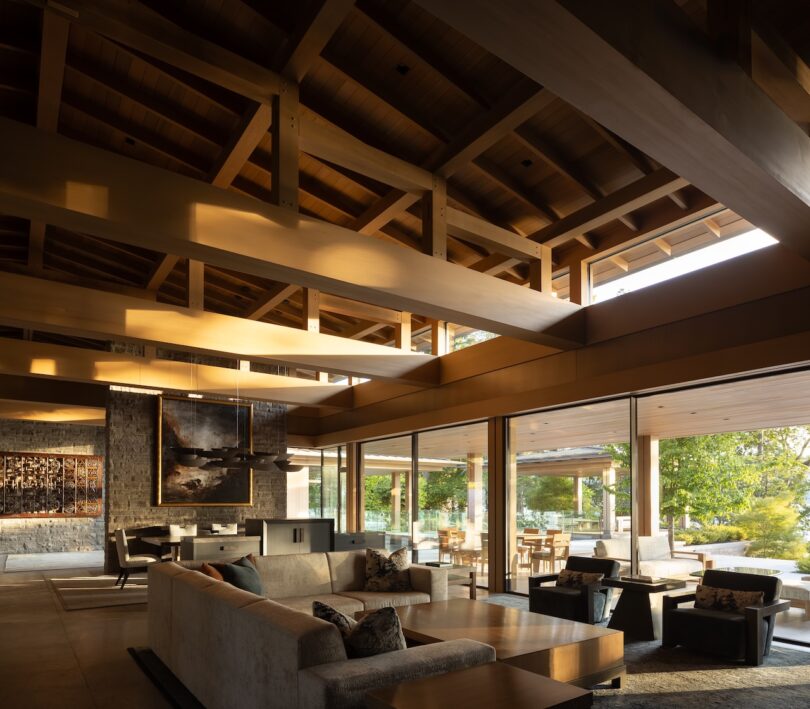 Modern living room with exposed wooden beams, large windows, neutral-toned furniture, and abundant natural light overlooking a green outdoor area.